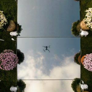 A drone's reflection seen on mirror surface surrounded by flowers and chairs outdoors.